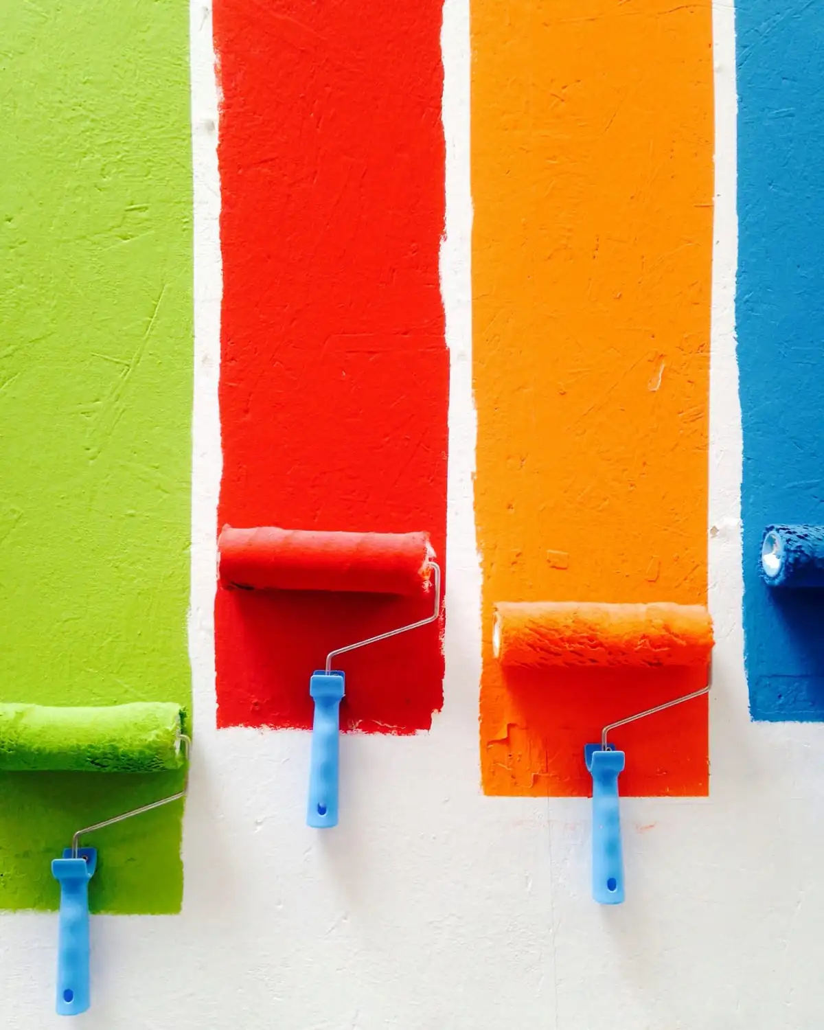 Four rollers laying stripes of green, red, orange and blue paint on a white wall