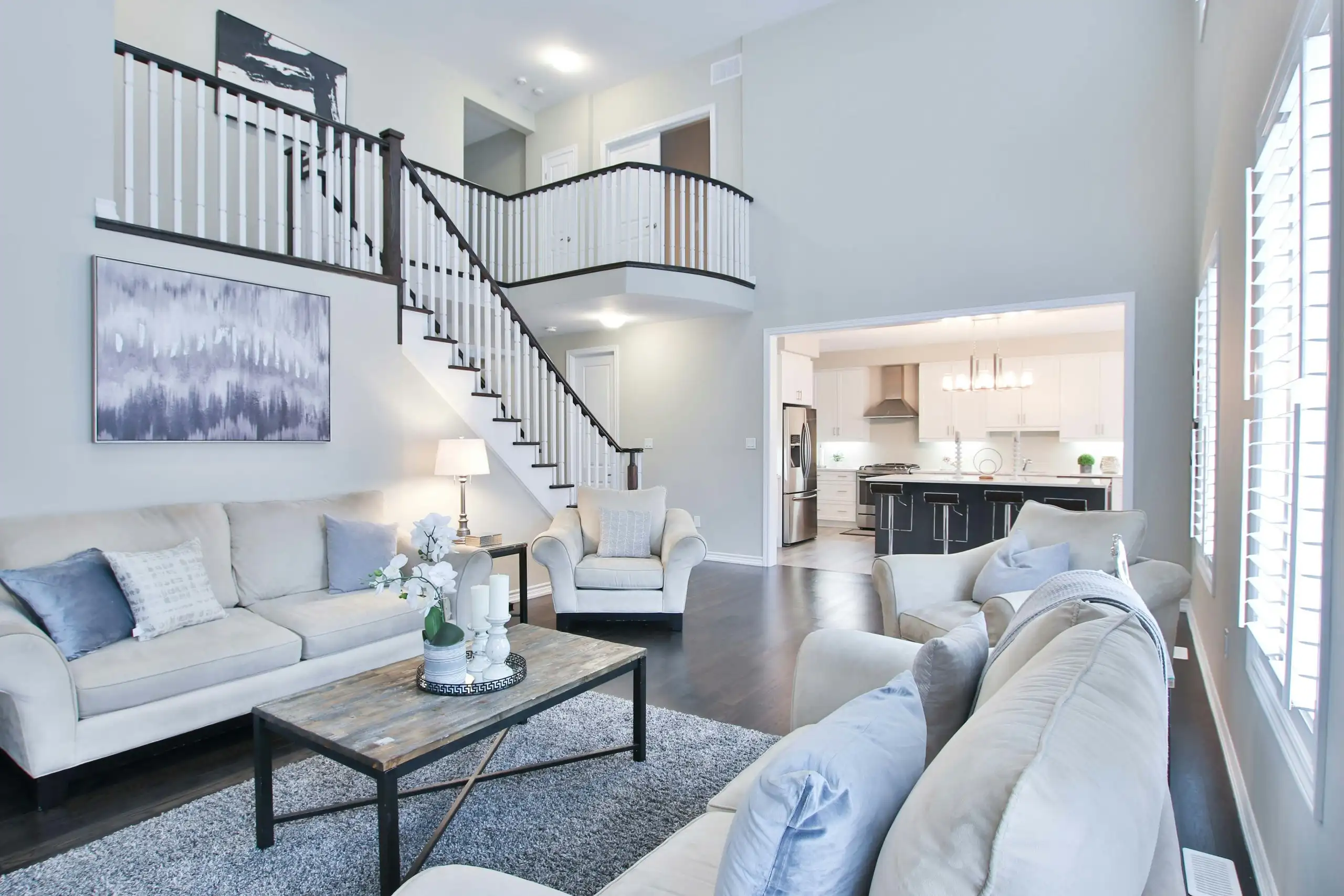 An open-plan living room with pale walls, a white staircase and grey sofas