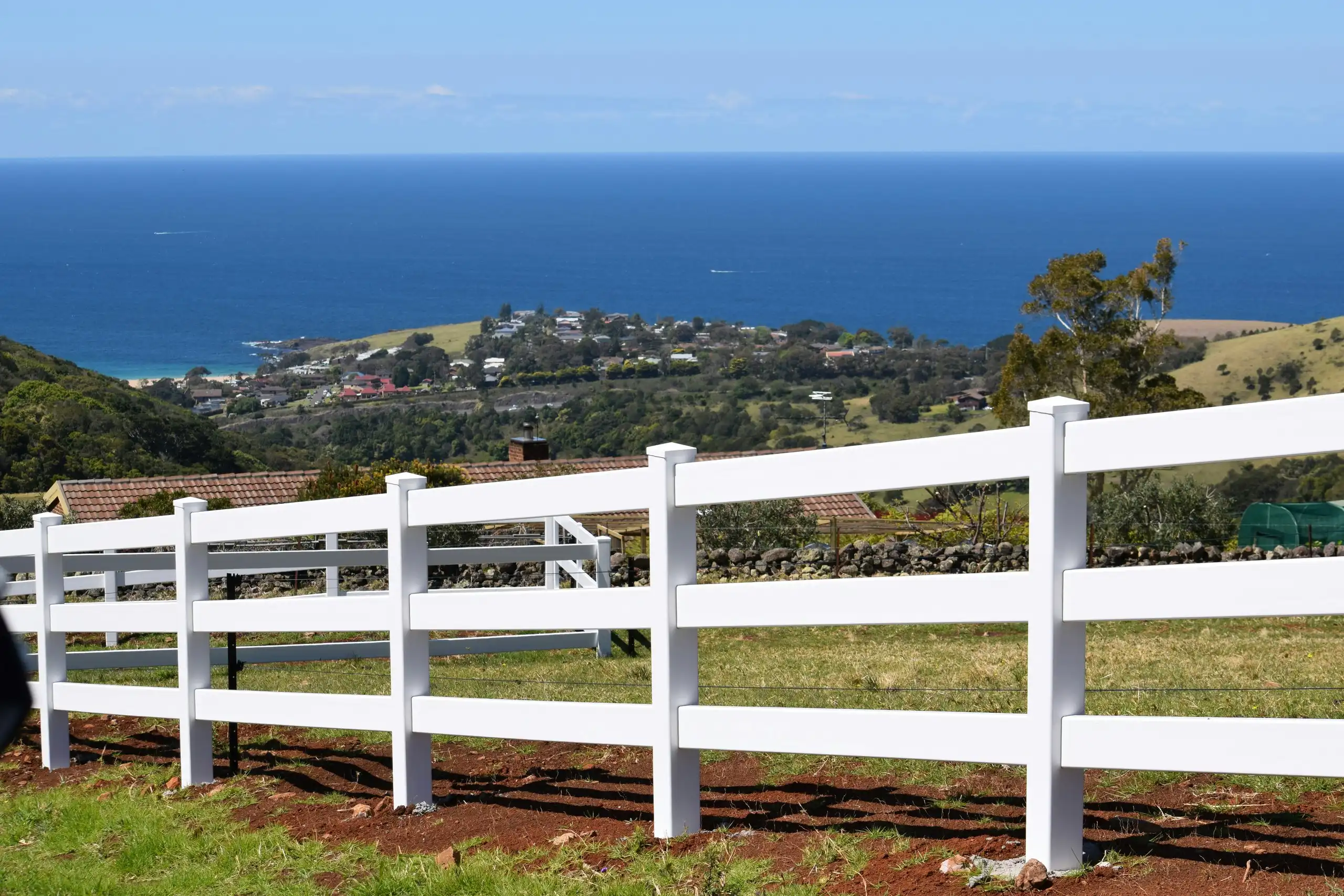 A white post-and-rail fence on a green slope above a coastal town and the sea
