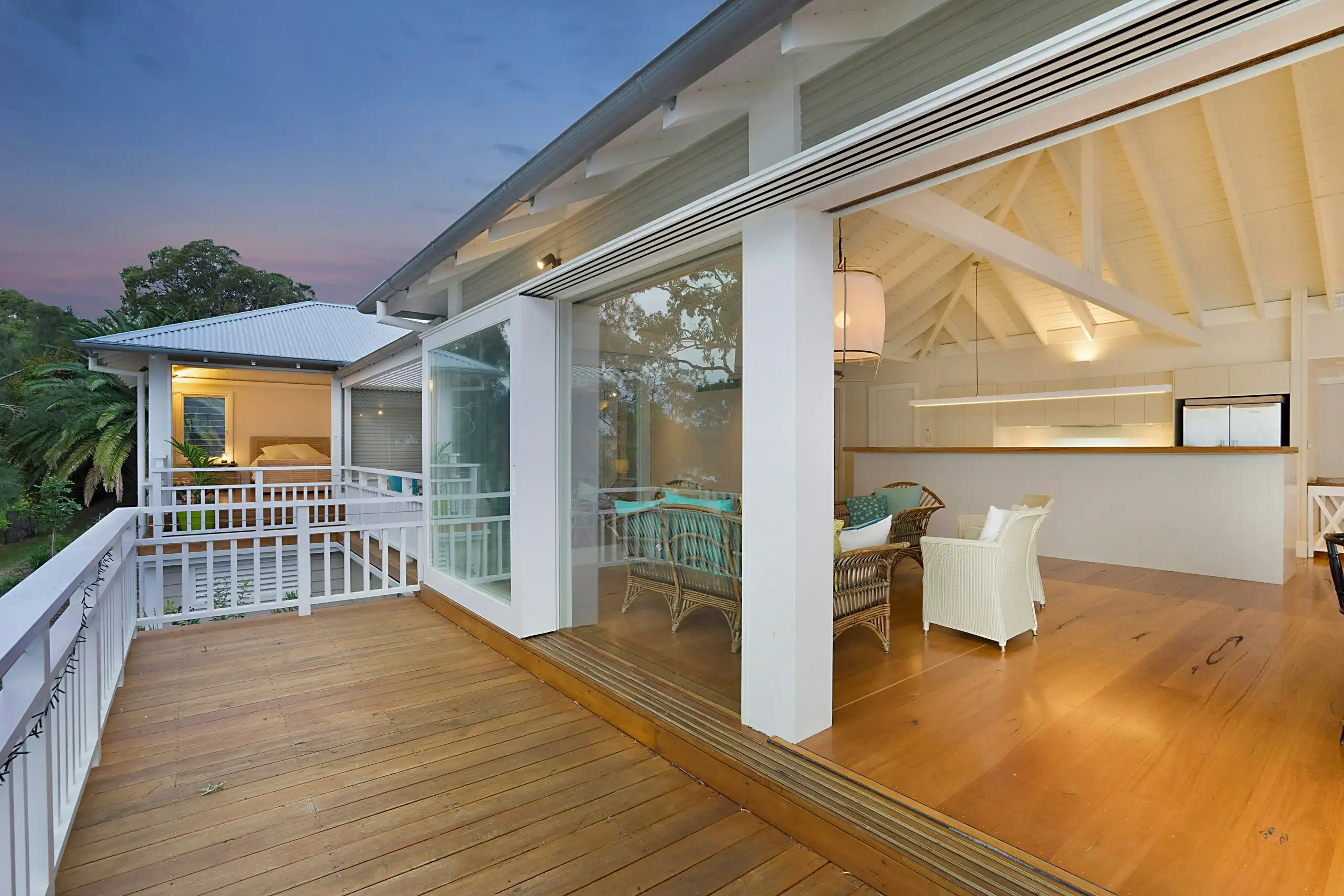 A timber deck on a raised weatherboard house at dusk, white balustrades and the lights on inside