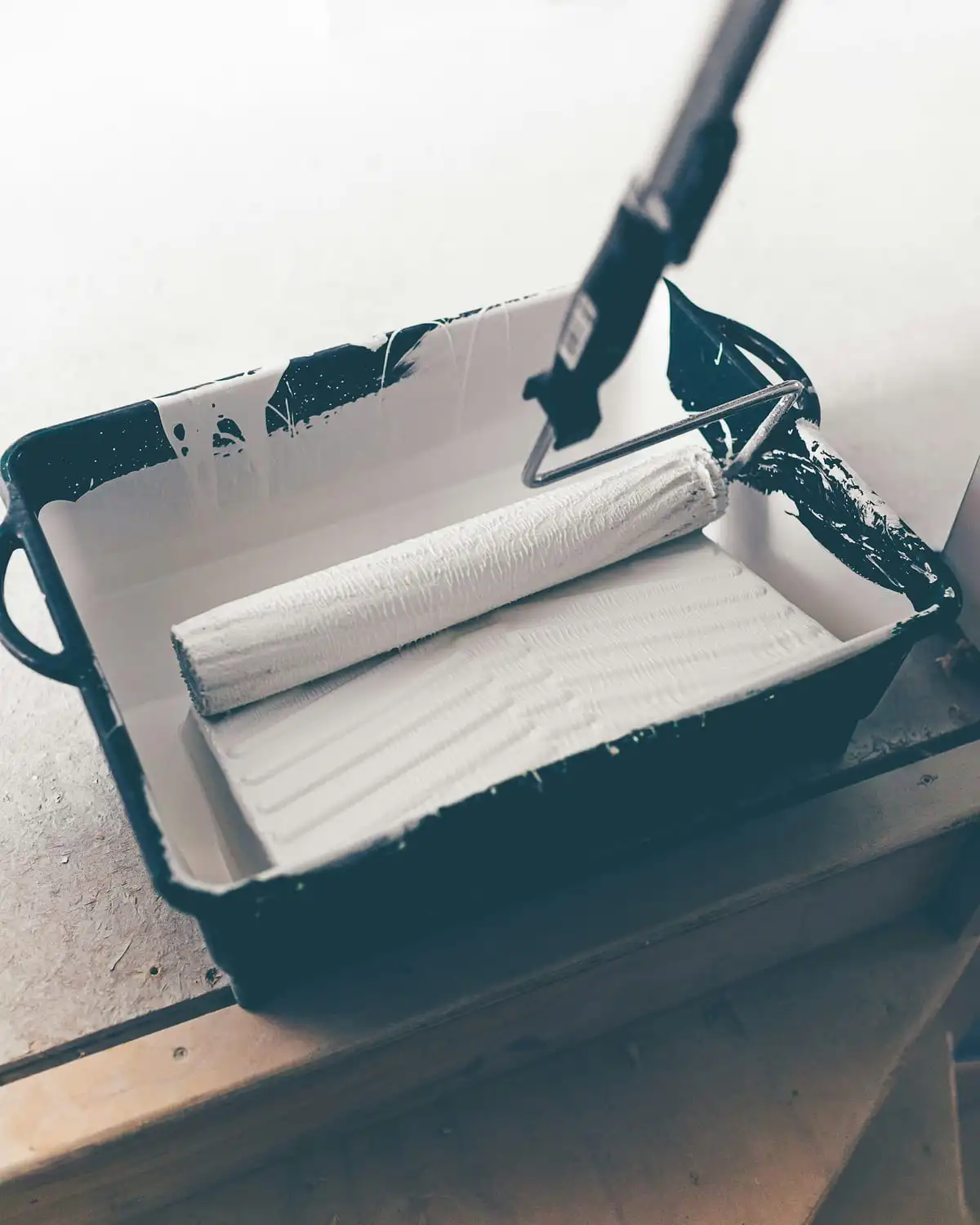 A paint roller resting in a tray, white paint on the roller and dark paint on the rim