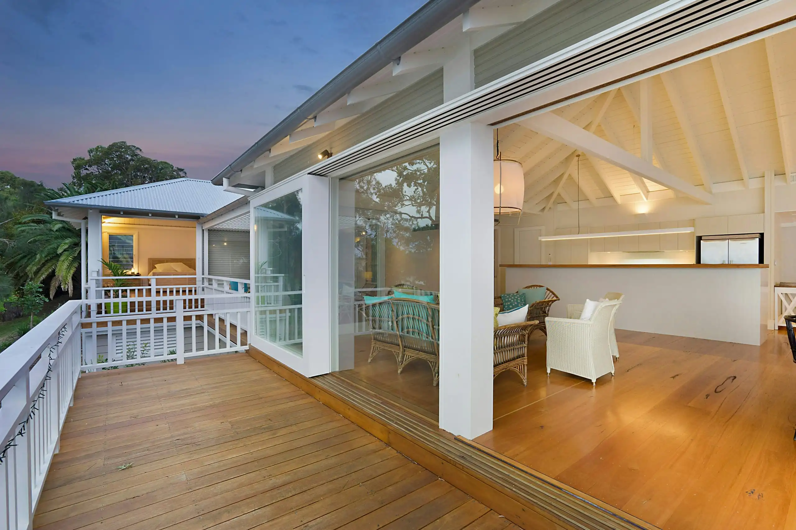 A timber deck on a raised weatherboard house at dusk, white balustrades and the lights on inside