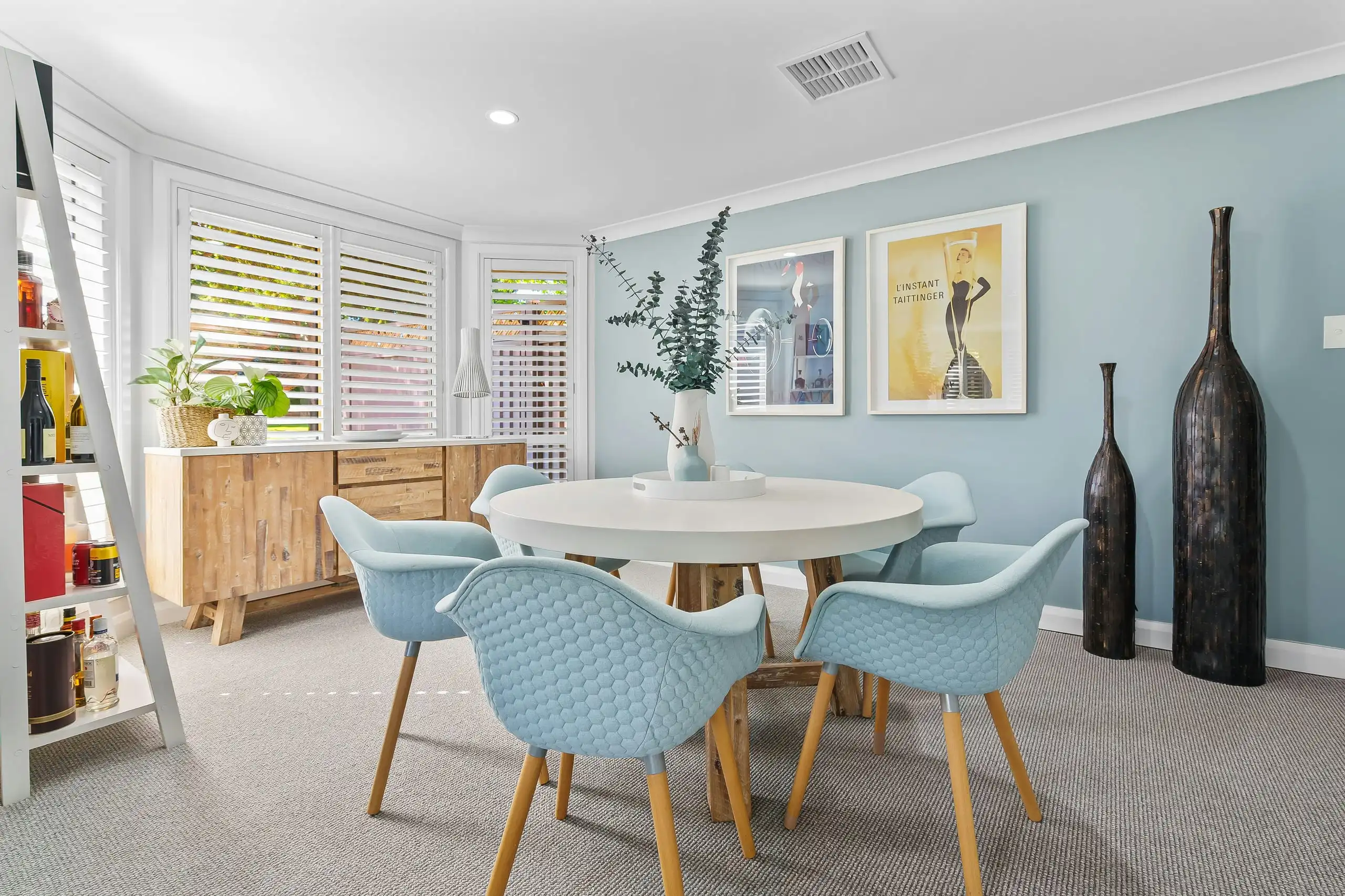 A dining nook with pale blue walls, white shutters, a round white table and blue chairs