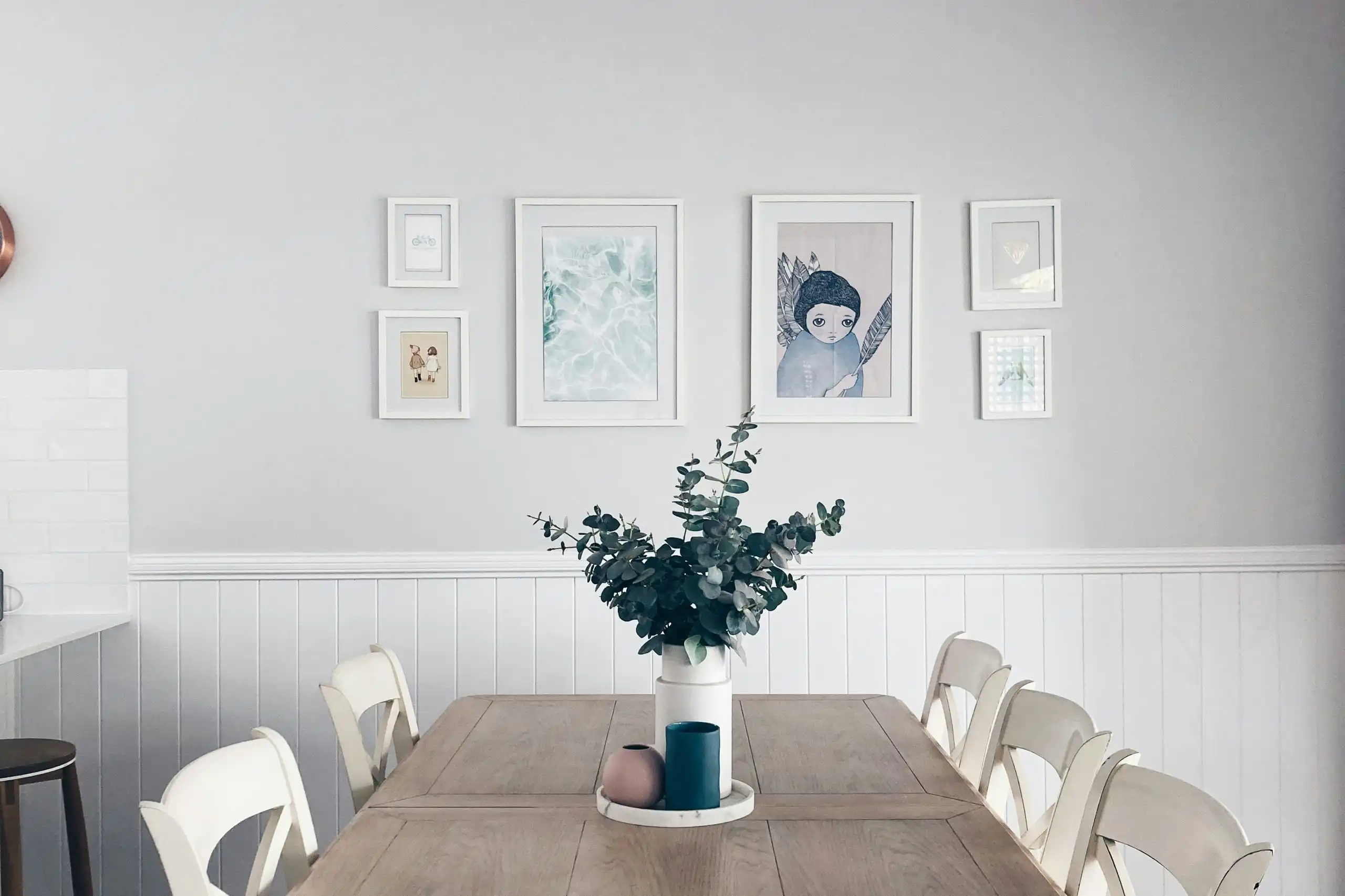 A dining table under a group of framed prints, white wainscoting below pale grey walls